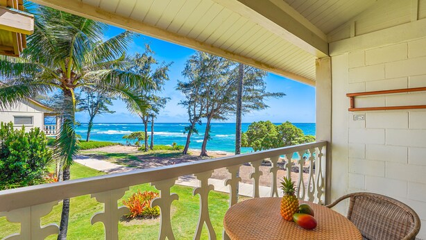 A balcony with a view of the ocean, a wicker table with fruits, and a white railing. - Ocean View Beachfront, Split AC Wifi, Pool Hot Tub (Kapaa)