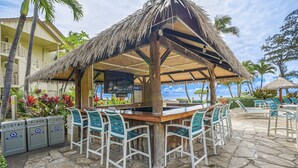 Un bar al aire libre con techo de paja y suelo de piedra, rodeado de sillas blancas y un área de lounge junto a la piscina con sillas y sombrillas azules.