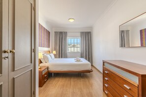 A hotel room with a bed, bedside table, dresser, window with curtains, and a mirror.