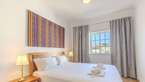 Et hotelværelse med en seng, sengeborde med lamper, et vindue med gardiner og et farverigt kunstværk på væggen.