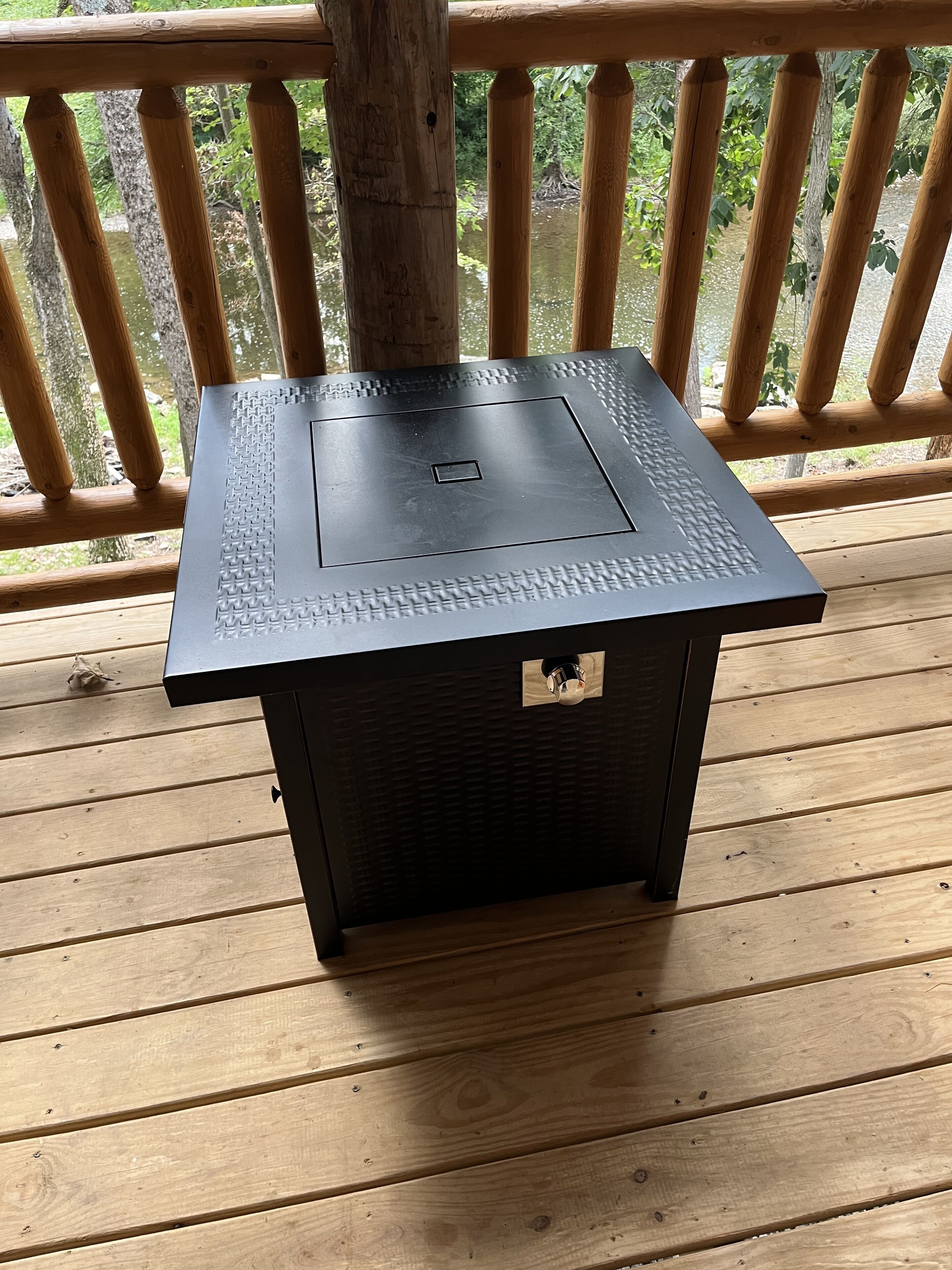 A square, black outdoor grill with a textured top, placed on a wooden deck with a railing in the background.