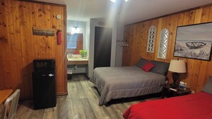 Una habitación con paneles de madera, una cama con ropa de cama gris, una colcha roja, una mesita de noche con una lámpara, un cuadro de un barco en la pared y un letrero que dice “BISHIN”.