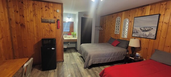 A room with wooden paneling, a bed with grey bedding, a red blanket, a nightstand with a lamp, a painting of a boat on the wall, and a sign that says "BISHIN".