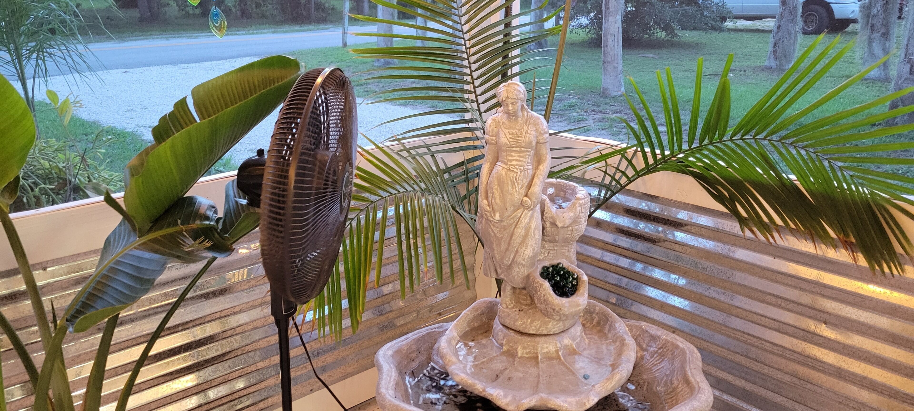 Una fuente de piedra con una estatua, rodeada de plantas tropicales y un ventilador de pie.