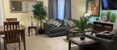 A living room with a sofa, two chairs, a coffee table, a ceiling fan, and a TV on the wall.