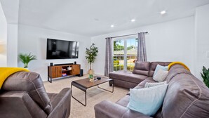 A modern living room with a brown leather sofa, a flat-screen TV, a wooden entertainment unit, and a large window with curtains.