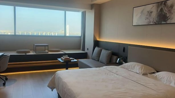 A modern hotel room with a large bed, a desk, a chair, a small table, and a large window offering a city view. - Jingdian Hotel (Nan'an)