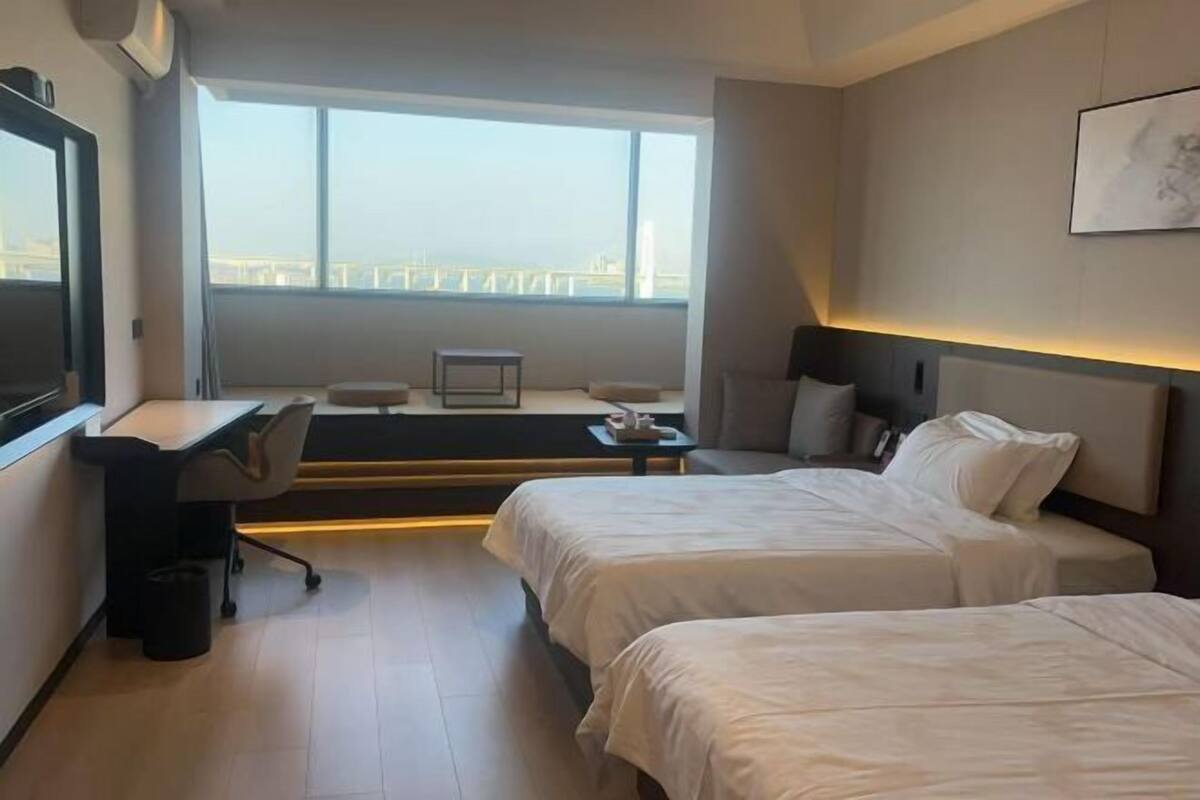 A hotel room with two beds, a desk, a chair, and a large window offering a city view.