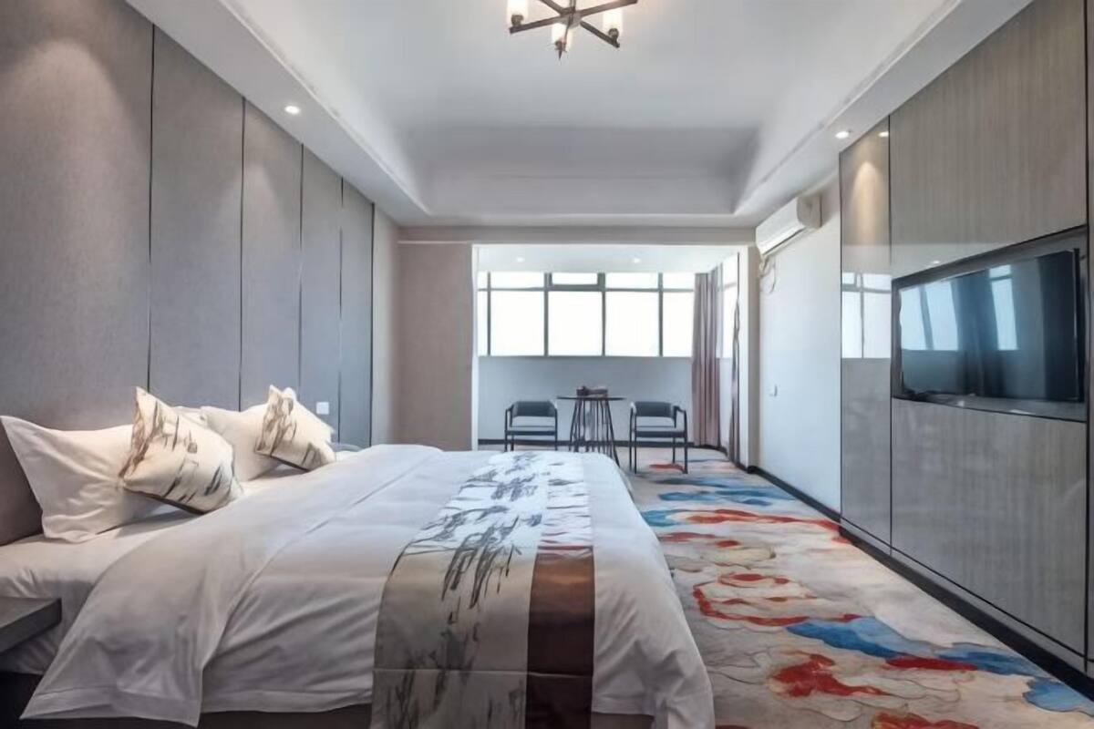 A modern hotel room with a large bed, a TV, a small table, and a view of the outdoors.