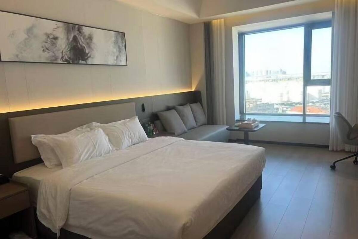 A hotel room with a large bed, a sofa, a small table, and a large window with a city view.