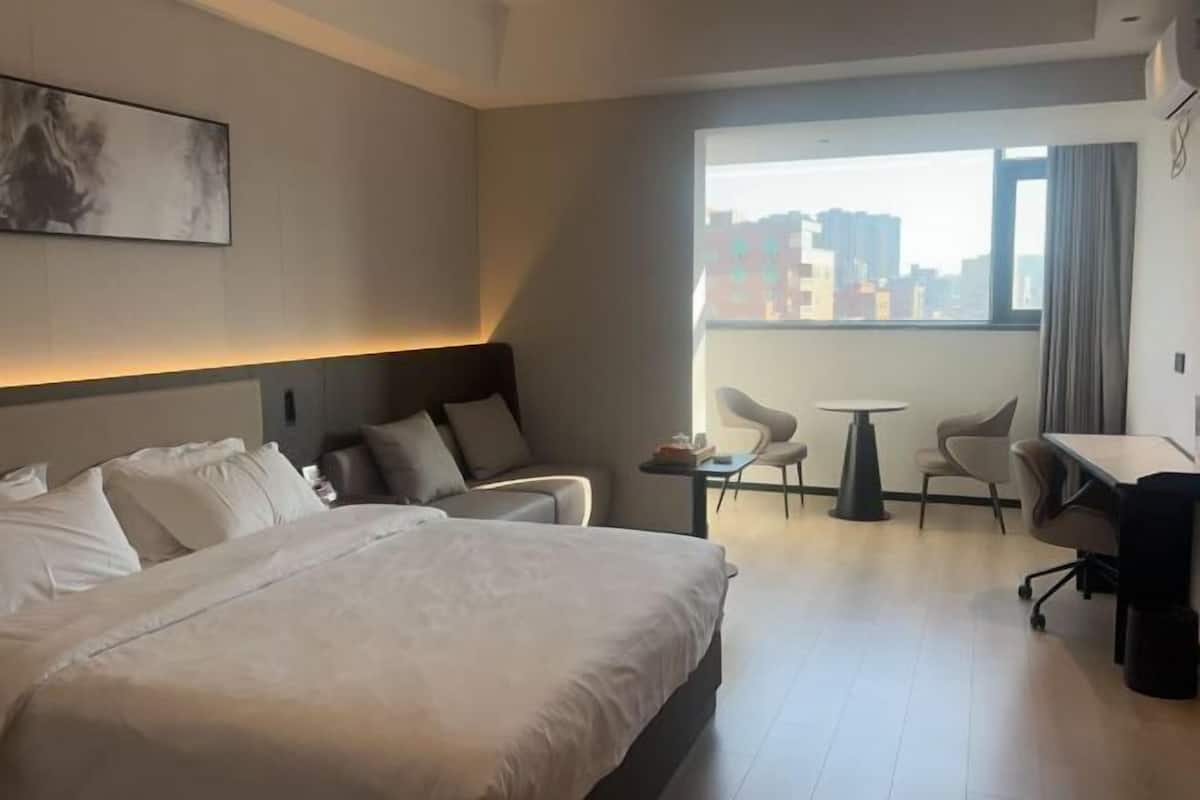 A hotel room with a bed, a desk, two chairs, a small table, and a large window with a city view.