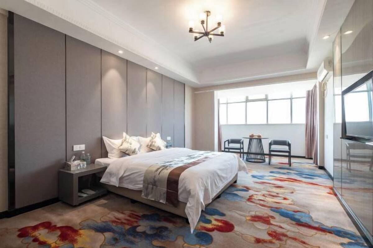 A hotel room with a large bed, a nightstand, a chair, a desk, and a large window.