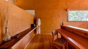 Un interior de madera con un banco, una silla, una mesita, un florero con plantas secas y una bata colgada en un perchero.