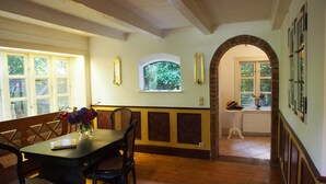 Un comedor con una mesa y sillas de madera, un jarrón con flores y un banco. Hay una ventana con vistas a árboles y un arco de ladrillo que conduce a otra habitación.