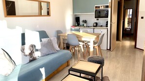 Ein kompakter Wohnbereich mit Sofa, Esstisch, Stühlen, einer kleinen Küchenzeile mit Mikrowelle und einer Wanduhr.