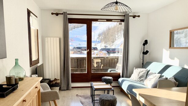 Ein gemütliches Wohnzimmer mit einem Holzesstisch, einer Couch und Blick auf eine verschneite Landschaft durch die Glastür.