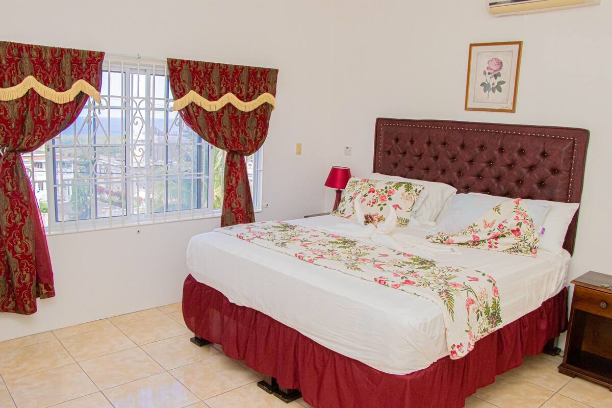 A bedroom with a large bed, red curtains, a window with a view, a bedside table, and a framed picture on the wall.