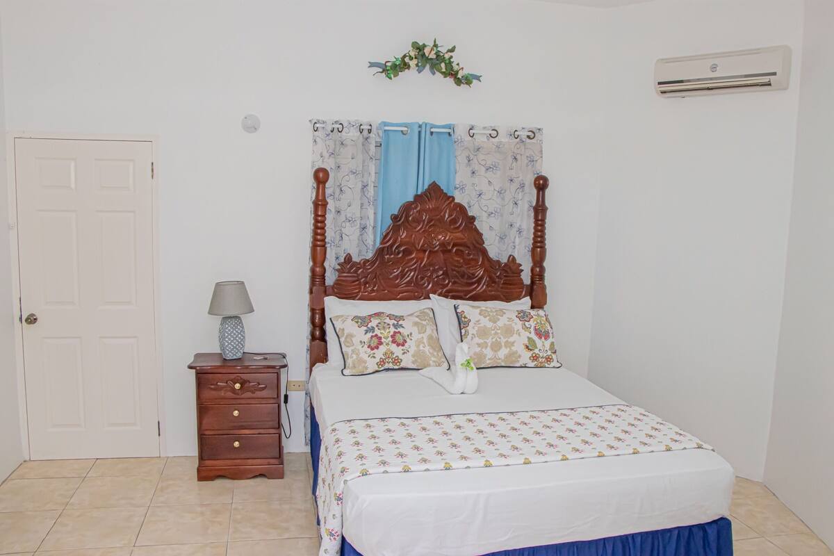 A bedroom with a wooden bed, a nightstand with a lamp, a white door, and a wall-mounted air conditioner.