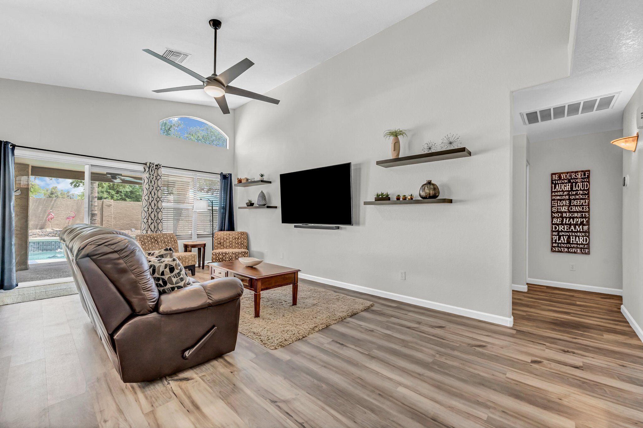 A modern living room with a brown leather sofa, a flat-screen TV, wooden flooring, and a ceiling fan.