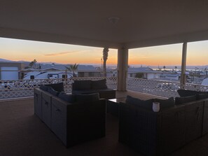 Ein überdachter Außenbereich mit Korbmöbeln und Blick auf die Stadtlandschaft bei Sonnenuntergang.