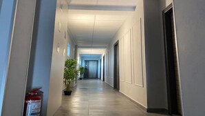 Un pasillo largo y estrecho con paredes de tonos claros y suelo de baldosas. Hay plantas en macetas a lo largo del pasillo, y se ve un extintor a la izquierda.