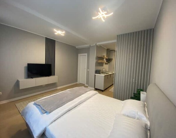 A modern hotel room with a large bed, a flat-screen TV mounted on the wall, a kitchenette, and a sliding door leading to another room.