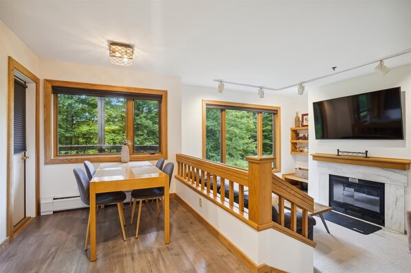 Un comedor con una mesa y sillas de madera, ventanas amplias con vista a árboles, una escalera con barandilla de madera y una chimenea con un televisor montado encima.