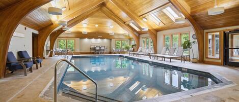 An indoor swimming pool with a wooden ceiling, lounge chairs, and a view of the outdoors.
