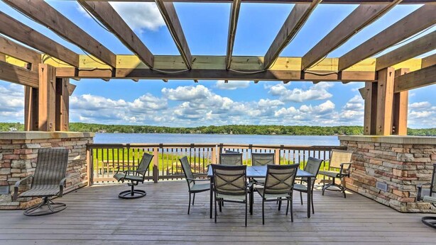 Une terrasse extérieure couverte, dotée de poutres en bois et d’un mur en pierre, avec une table et des chaises, et offrant une vue sur un lac.