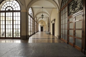 A long corridor with high ceilings, large arched windows, and a polished stone floor.