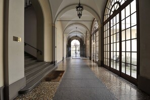 A long, arched hallway with a tiled floor, a staircase on the left, and large windows on the right.