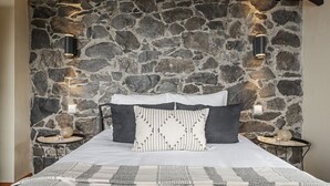 Ein Schlafzimmer mit einer Steinwand, einem Bett mit weißer und schwarzer Bettwäsche, zwei Nachttischen mit Lampen und einem gemusterten Teppich.