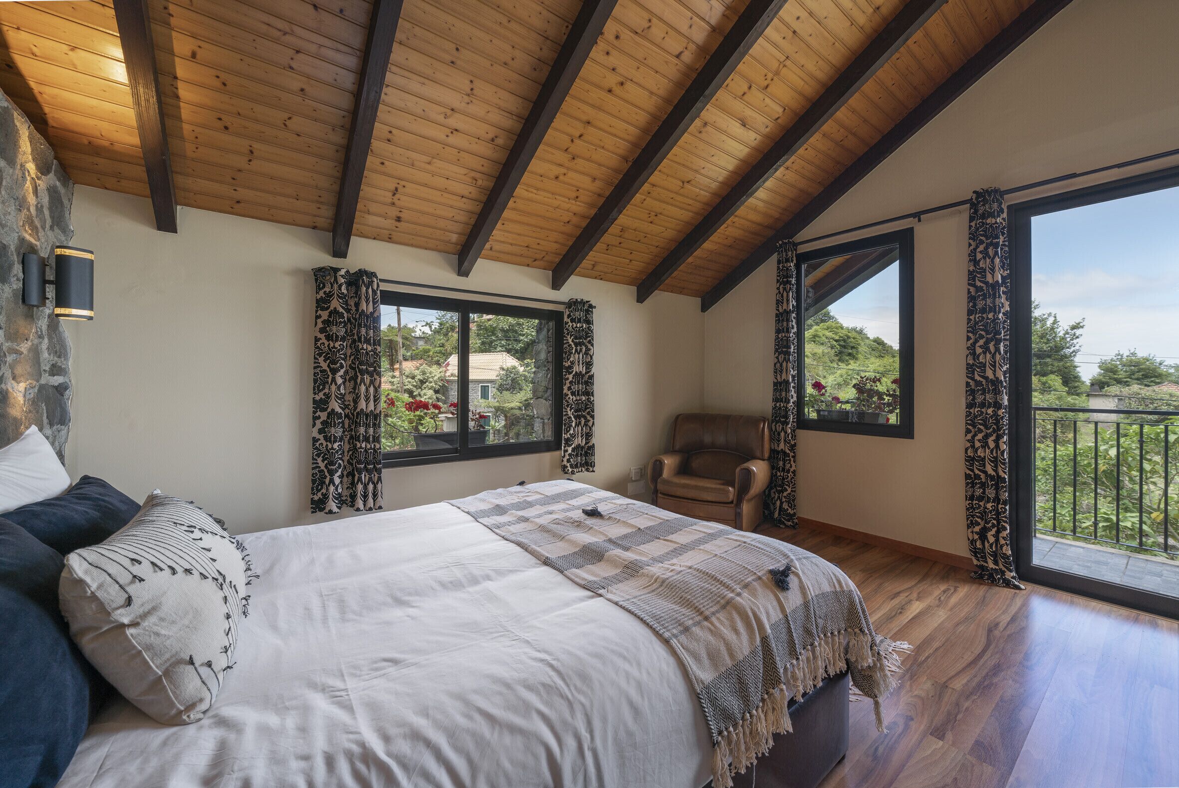 A bedroom with a large bed, wooden ceiling beams, a stone wall, and two windows with patterned curtains.