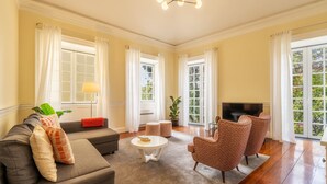Una sala de estar con sofá, sillones, una mesita de centro y televisor. Amplias ventanas con cortinas ofrecen vistas a zonas verdes.