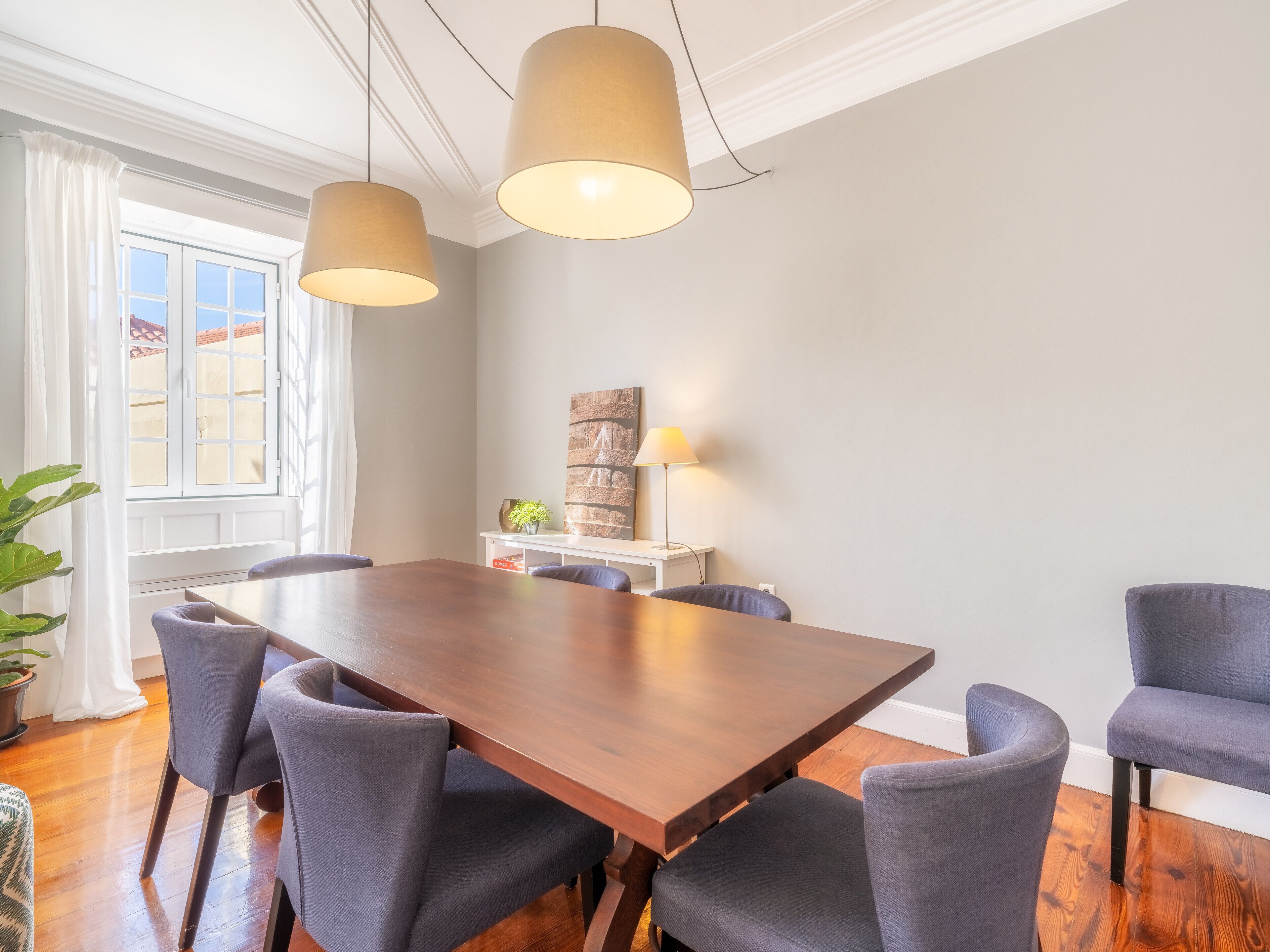 A dining area with a wooden table and six chairs, two pendant lights, a potted plant, and a window with curtains.