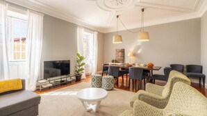 Una sala de estar moderna con un sofá gris, una mesa de centro blanca y otomanas estampadas. Hay un comedor con una mesa y sillas, y un televisor en una esquina.