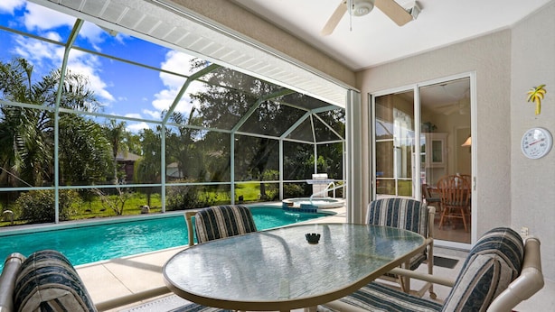 Uma área externa coberta com mesa e cadeiras de vidro, piscina e vista para o quintal.