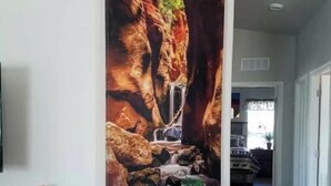 A living room with a large painting of a waterfall, a wooden cabinet with drawers, and a small table with a plant. - Zion Fence Line - Walk to Trailhead  - Secluded (Orderville)
