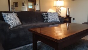 A living room with a dark-colored sofa, patterned cushions, a wooden coffee table, a wall clock, a lamp, and a framed picture. - Quiet Barn Loft at Rock'n'Rose (Morganton)