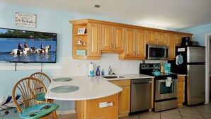 A kitchen with wooden cabinets, stainless steel appliances, a dining table with chairs, and a wall-mounted television displaying a beach scene.