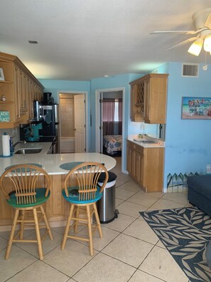 Cocina compacta con armarios de madera, una isla central con sillas, un cubo de basura y un tapete estampado.