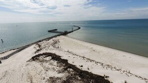 Vue aérienne d’une plage avec une jetée en bois s’étendant dans l’eau, un ciel dégagé et une mer calme.