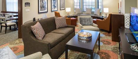 A hotel lobby with a brown sofa, armchairs, a coffee table, and a television. There are framed pictures on the wall and a dining area in the background.