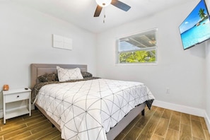 Quarto com cama, mesa de cabeceira, ventilador de teto, televisão na parede e janela com persianas.