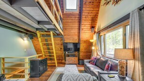 Een gezellig cabin interieur met een slaapkamer op entresol, een open haard, een televisie en een groot raam met gordijnen.