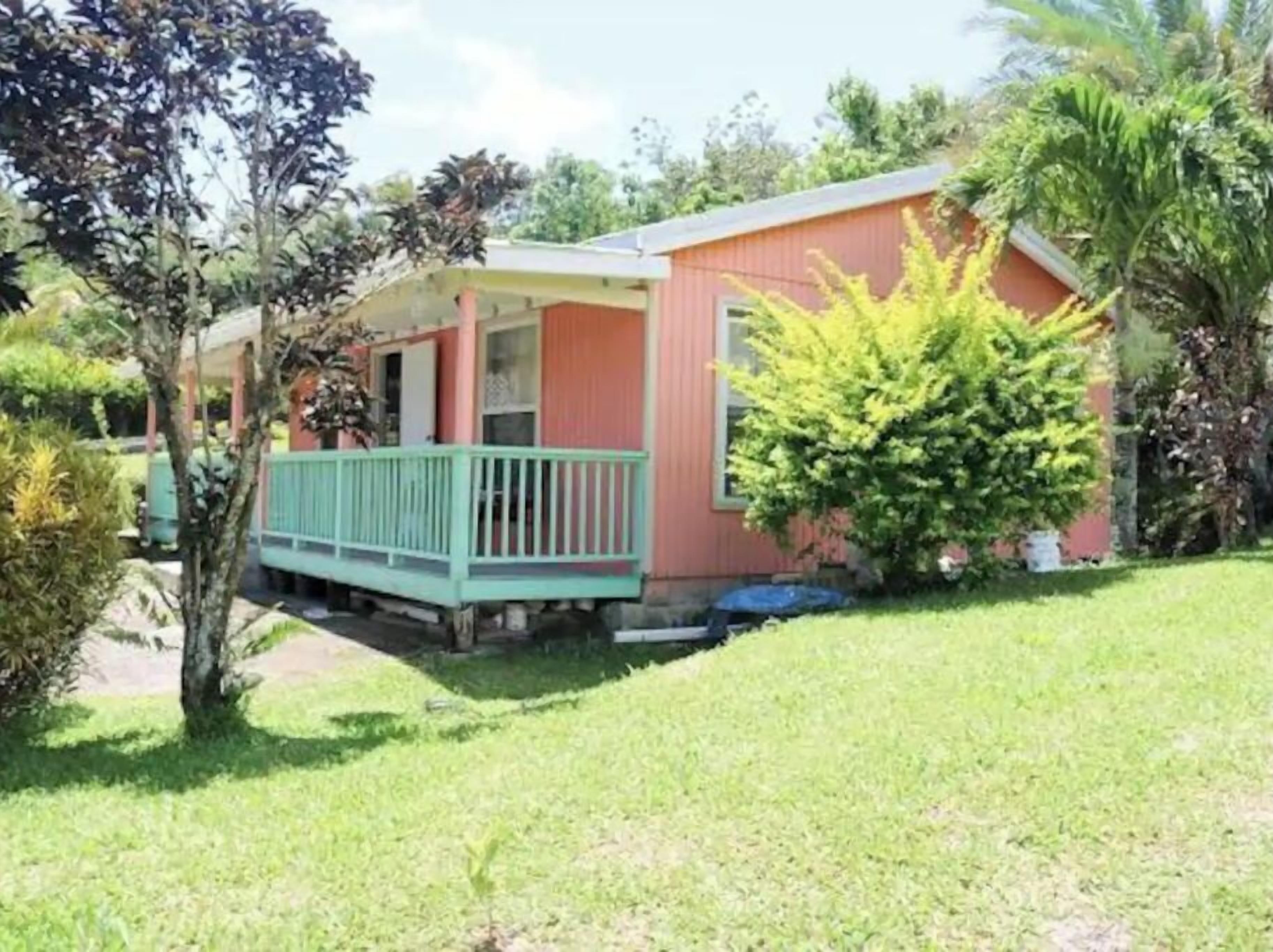 A small, single-story house with a porch, surrounded by green grass and trees.