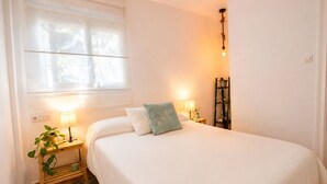 Een netjes opgemaakt bed met twee kussens, een nachtkastje met een lamp, een kleine plant en een raam met zonwering.