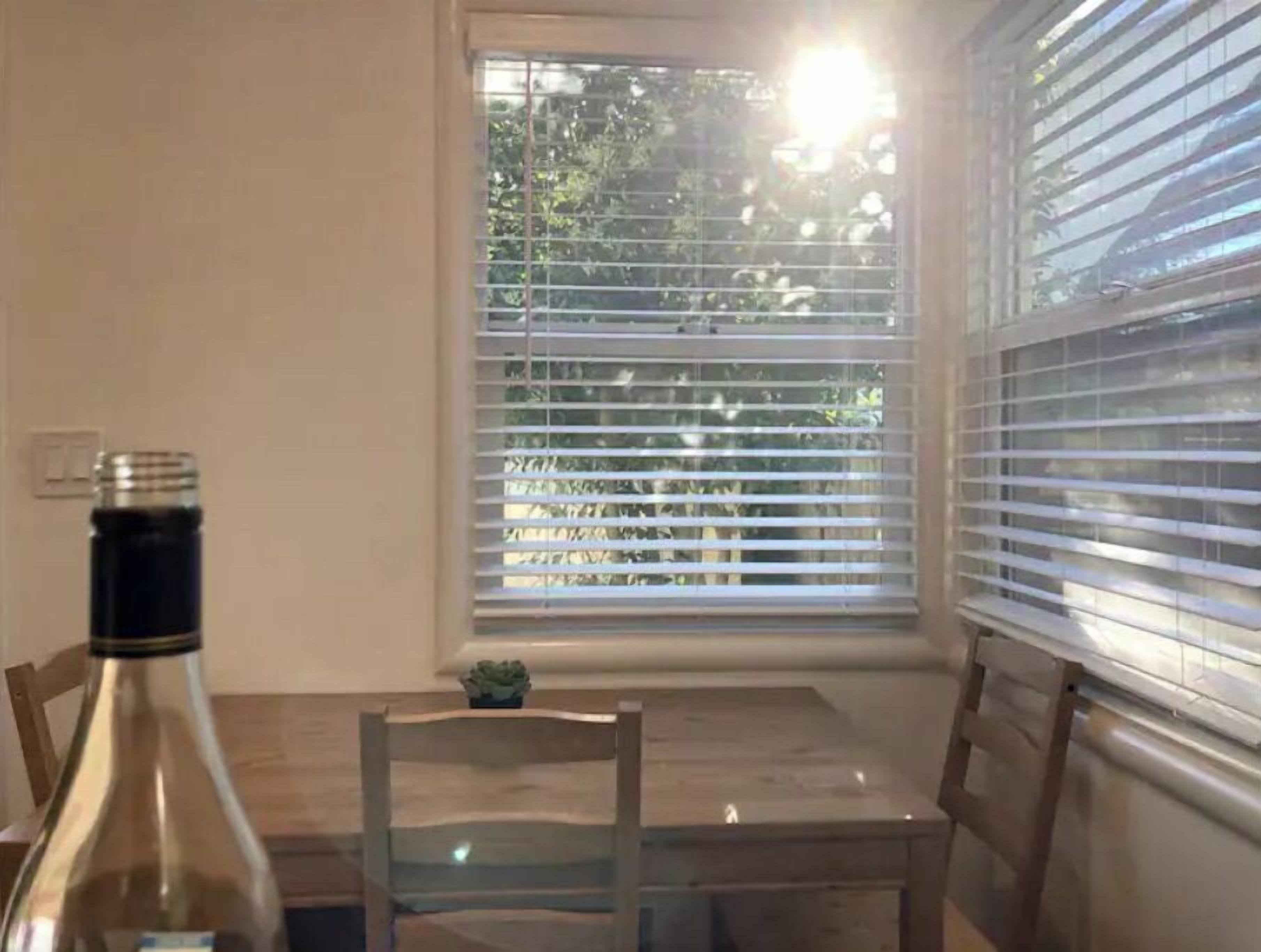 Un coin repas avec une table et des chaises, une bouteille posée sur la table et une fenêtre avec des stores.
