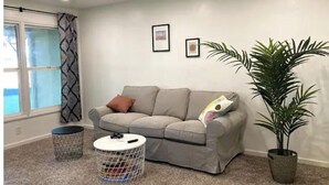 Una sala de estar con un sofá gris, dos cojines, una mesita redonda con superficie blanca, una planta en maceta y una ventana con cortinas estampadas.