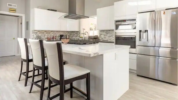Una cocina moderna con armarios blancos, una isla grande con taburetes de bar, un refrigerador de acero inoxidable y un horno.